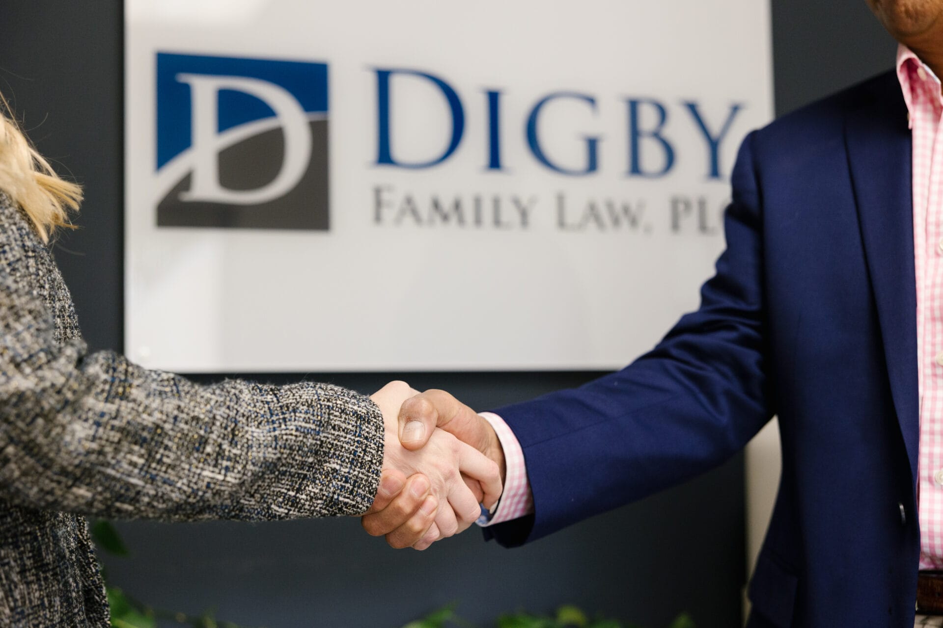 A close-up of a handshake between two people in a professional setting. One person wears a textured blazer, and the other wears a navy suit jacket with a pink shirt. In the background, a sign reads "Digby Family Law, PLLC," indicating a legal or business agreement.