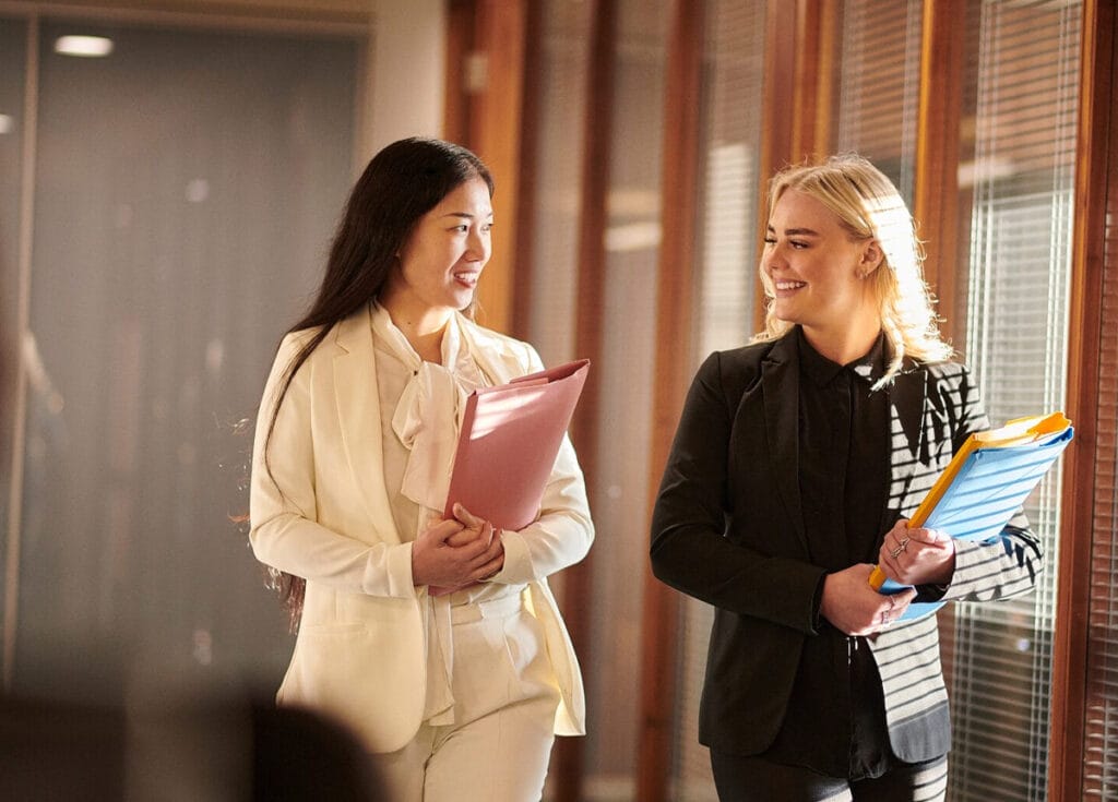Two professional women walk together down a sunlit office corridor. One wears a white suit and holds a pink folder, while the other wears a black suit and carries blue and yellow file folders. They are smiling and engaged in conversation, creating a friendly and collaborative atmosphere.