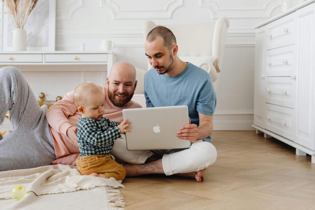 Same sex couple - two men hold a laptop in their lap while their nearby baby touches the laptop curiously