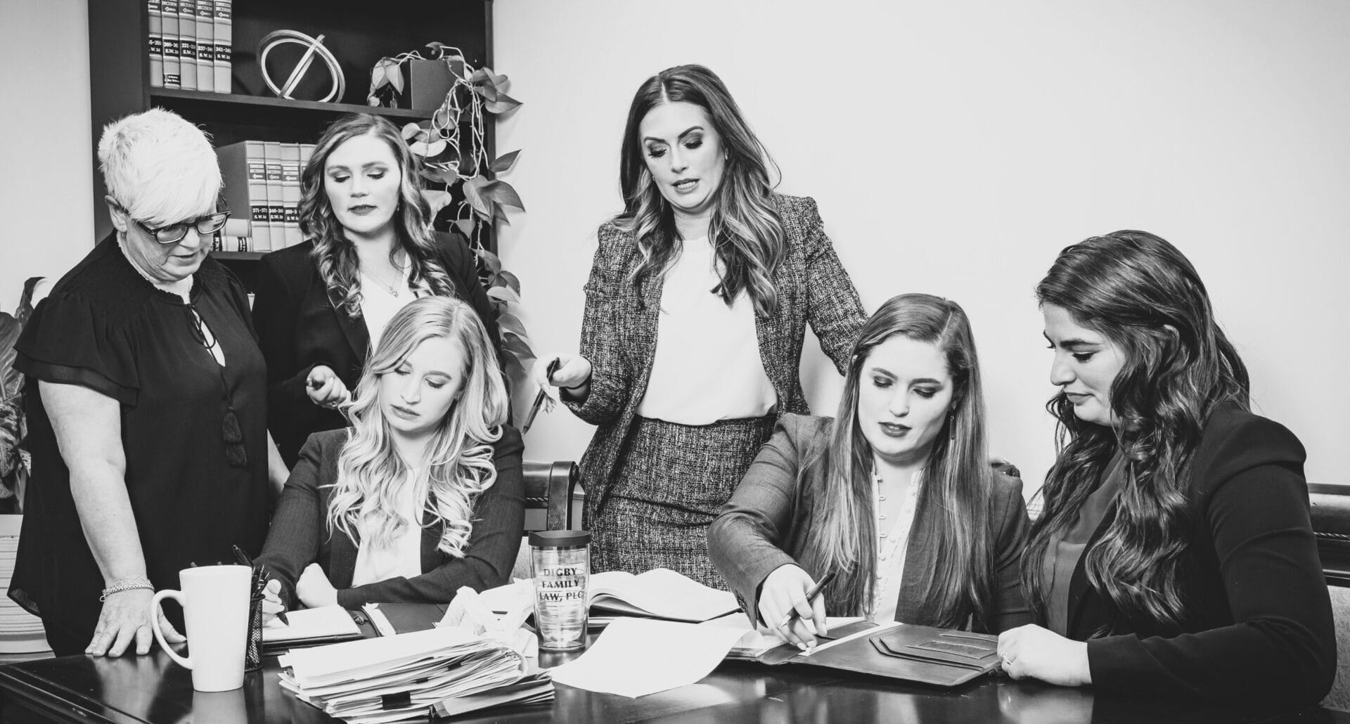 A group of six women gather around a desk in a professional setting. Some are seated and reviewing documents, while others stand and discuss. Bookshelves and decor are visible in the background, giving the scene a formal office atmosphere.