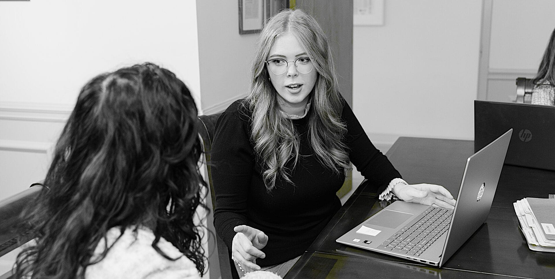 A professional woman with long blonde hair and glasses sits at a desk, engaged in conversation with another woman with curly dark hair. A laptop and papers are visible on the table, emphasizing a collaborative work environment.