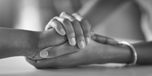 Two hands gently clasp each other in an intimate gesture of support and empathy. The black-and-white image conveys a sense of connection and care.