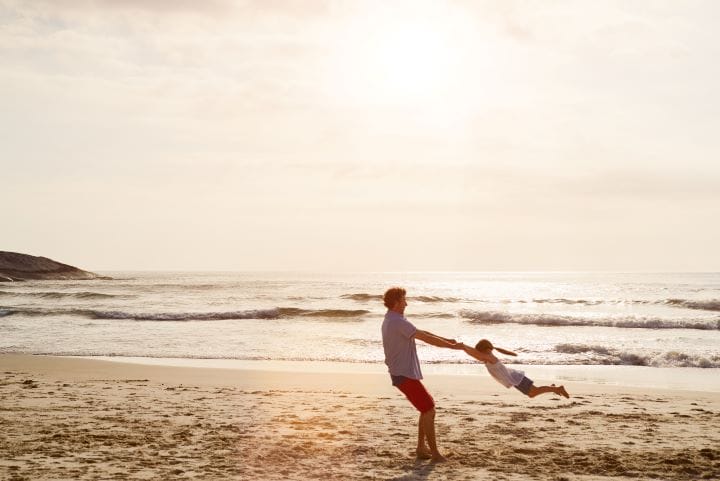 Alt text: A man in red shorts and a white shirt spins a young child in the air by holding their hands on a sandy beach. The ocean waves and a serene sky with soft sunlight fill the background, creating a warm and joyful atmosphere.