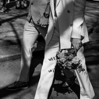 A black-and-white photo of a couple walking hand-in-hand. One person wears a tailored white suit and holds a bouquet of flowers, while the other wears a light-colored suit and a watch. Shadows of tree branches cast patterns on the ground, adding depth and texture to the scene.