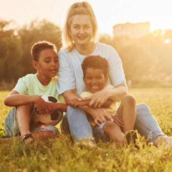 Adult woman sits with two black kids on the field at summer daytime.