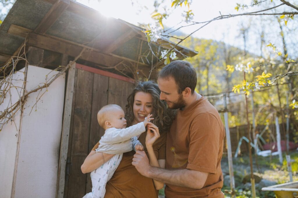 A young family stands outdoors on a sunny day, surrounded by greenery. A smiling woman with curly hair holds a baby, while a man in an orange shirt gently touches the baby's hand. The setting includes a rustic wooden shed and blossoming branches, creating a warm, natural scene.