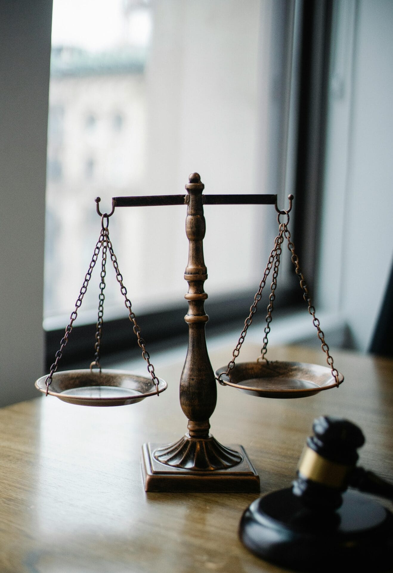 A close-up of a bronze balance scale sitting on a wooden desk, symbolizing justice and fairness. In the foreground, a judge’s gavel is partially visible, and a blurred window with an urban view forms the background.